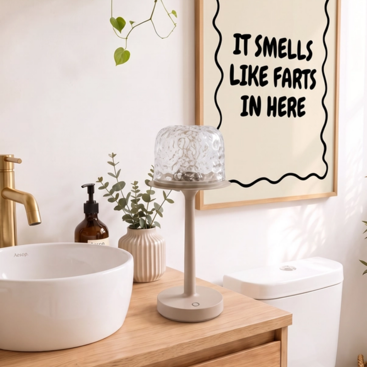 A framed wall art print with the text 'IT SMELLS LIKE FARTS IN HERE' in black lettering on a cream background, hanging in a bright, modern bathroom above a wooden vanity with a white vessel sink and a glass candle holder.